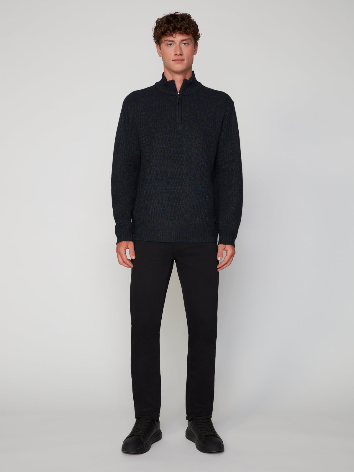 A young man with curly brown hair faces forward, wearing a dark PROJEK RAW MEN’S 1/4 ZIP MOCK NECK PULLOVER SWEATER, black pants, and black shoes against a plain light gray background.