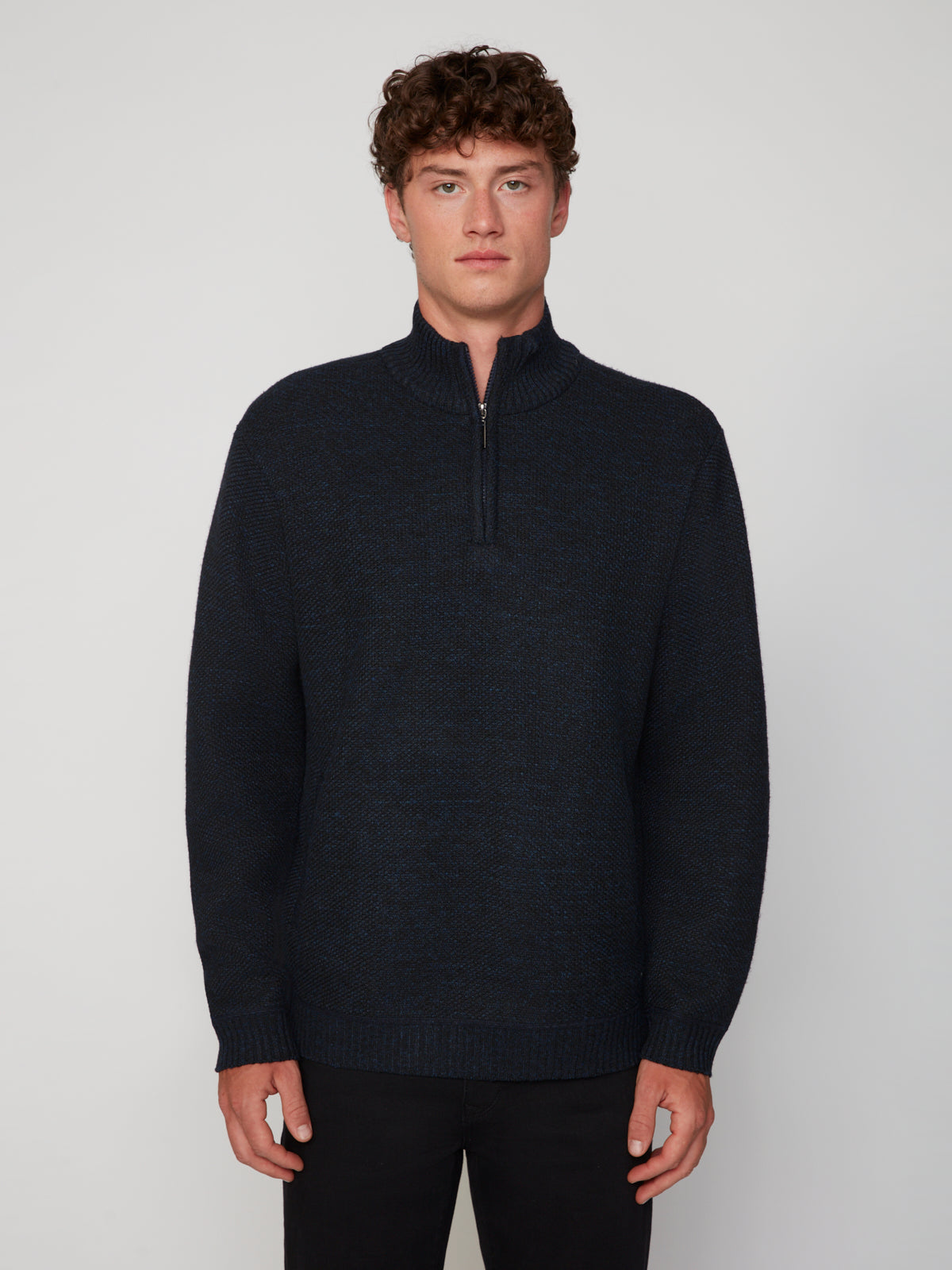 A young man with curly brown hair models the PROJEK RAW MEN’S 1/4 ZIP MOCK NECK PULLOVER SWEATER in dark blue and black pants, standing against a light gray background and facing the camera with a neutral expression.
