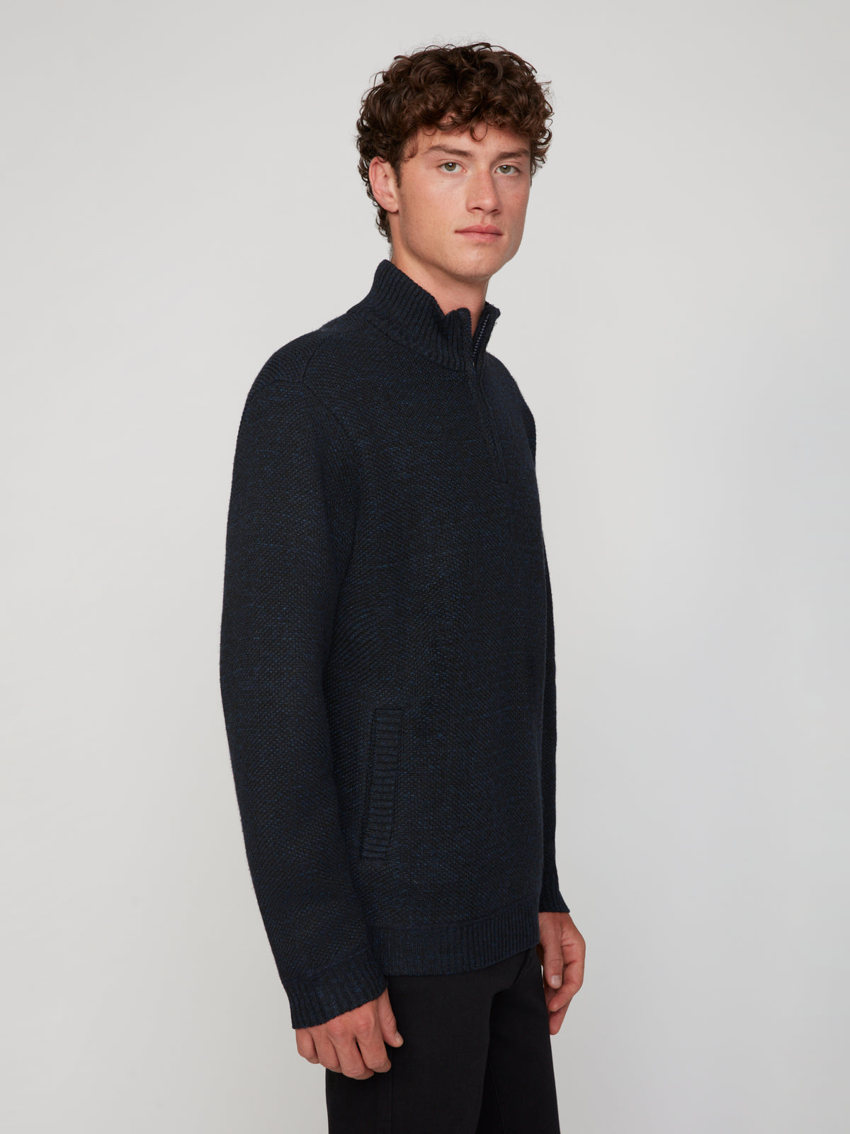 A young man with curly brown hair wears a dark blue PROJEK RAW MEN’S 1/4 ZIP MOCK NECK PULLOVER SWEATER with side pockets, standing against a plain light gray background and facing slightly to the side.