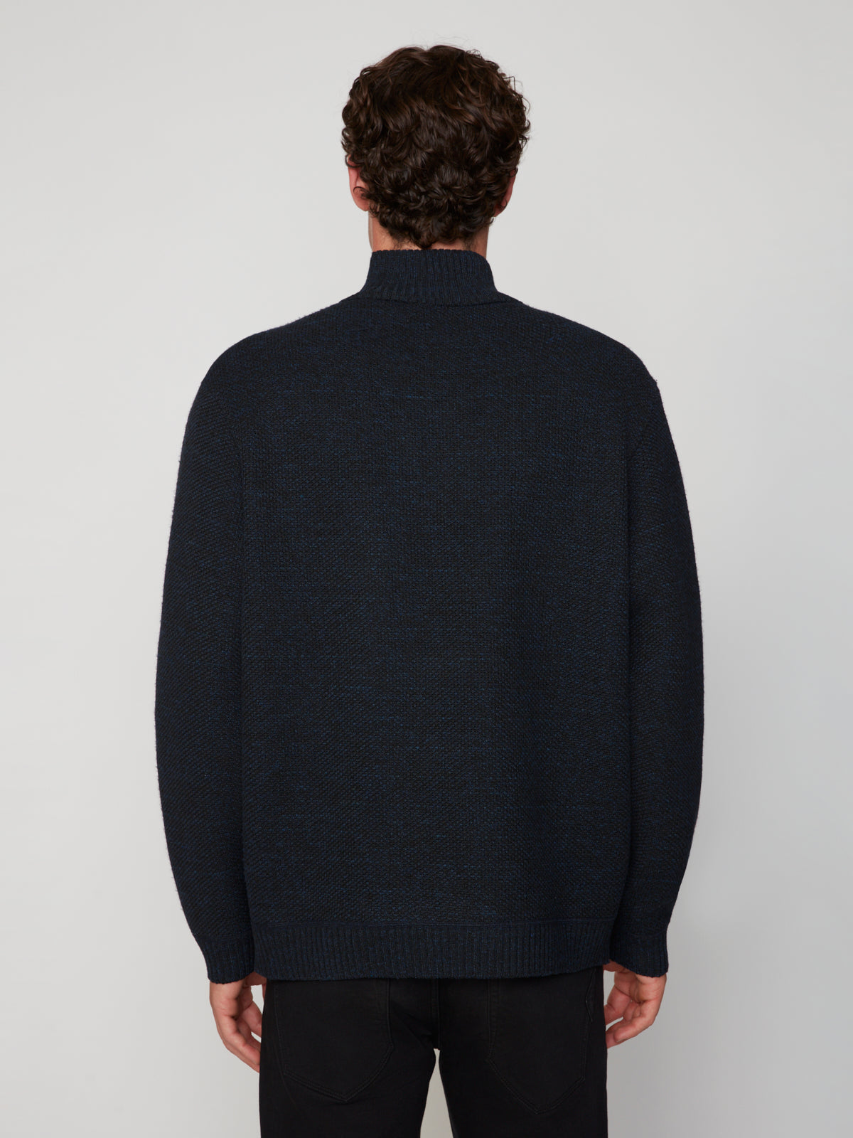 A person with short, curly brown hair is seen from the back wearing a PROJEK RAW MEN’S 1/4 ZIP MOCK NECK PULLOVER SWEATER in dark navy and black pants, standing against a plain light gray background.
