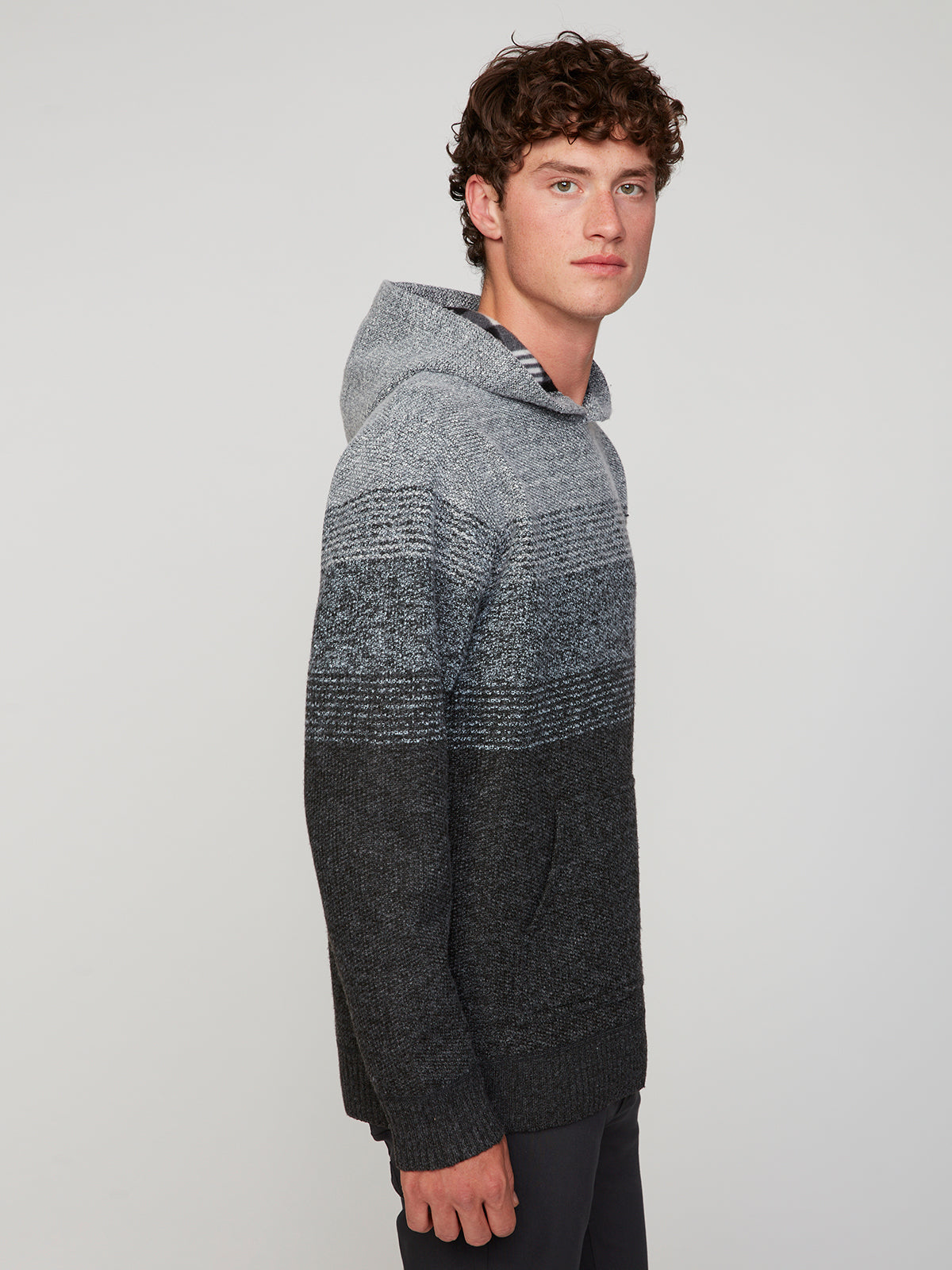A young man with curly brown hair models the PROJEK RAW MENS KNIT HOODED SWEATER BONDED WITH POLAR FLEECE, featuring a gray and black gradient. He stands sideways, facing the camera against a plain light background.