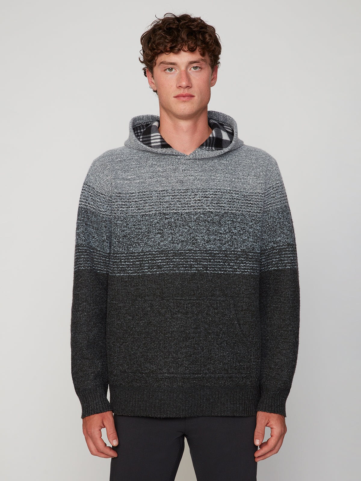 A young man with curly brown hair wears the PROJEK RAW MENS KNIT HOODED SWEATER BONDED WITH POLAR FLEECE in gradient grey and black, standing against a plain light background and looking directly at the camera.