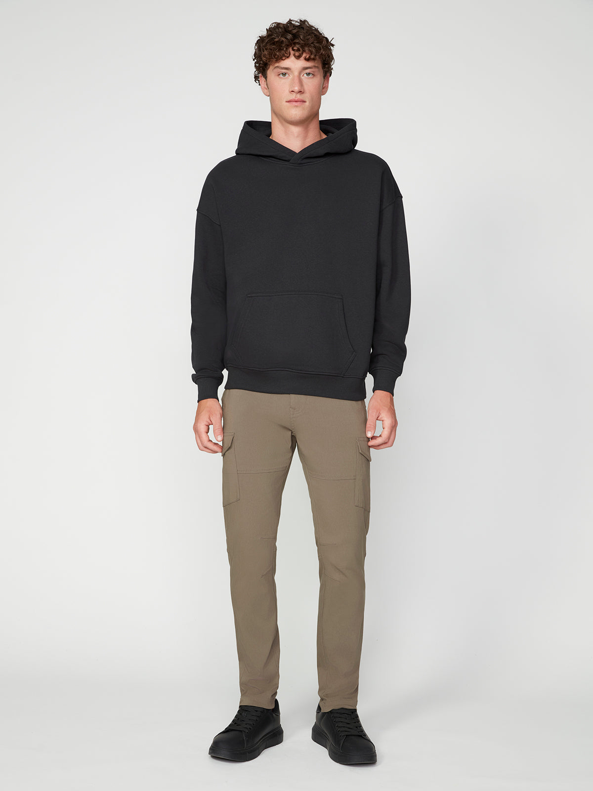 A young man with curly hair faces forward, wearing a PROJEK RAW MENS LONG SLEEVE CARBON PEACHED FLEECE HOODIE, khaki cargo pants, and black sneakers against a plain background.