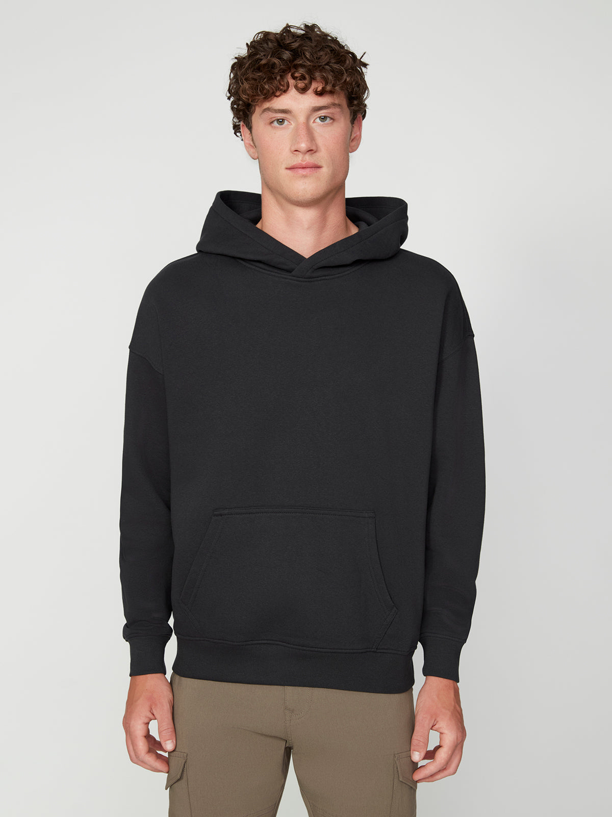 A young man with curly brown hair models the PROJEK RAW MENS LONG SLEEVE CARBON PEACHED FLEECE HOODIE in black and beige pants, standing against a light gray background with a neutral expression and relaxed hands by his sides.