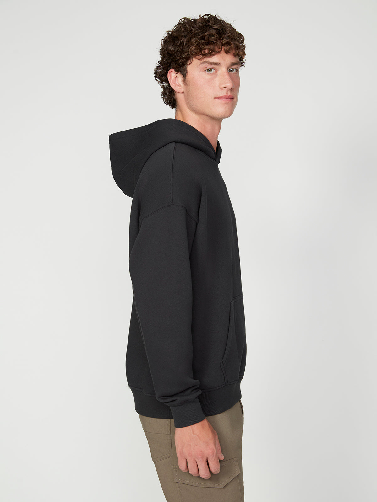 A young man with curly brown hair wears the PROJEK RAW MENS LONG SLEEVE CARBON PEACHED FLEECE HOODIE in black and light brown pants, standing sideways and looking at the camera against a plain white background.