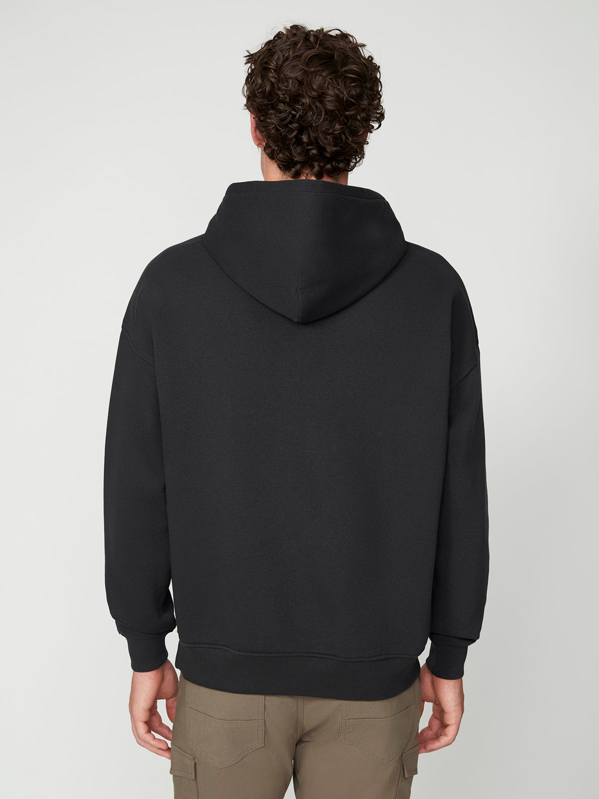 A person with curly brown hair is seen from the back, wearing an oversize fit PROJEK RAW MENS LONG SLEEVE CARBON PEACHED FLEECE HOODIE and khaki cargo pants, standing against a plain light background.