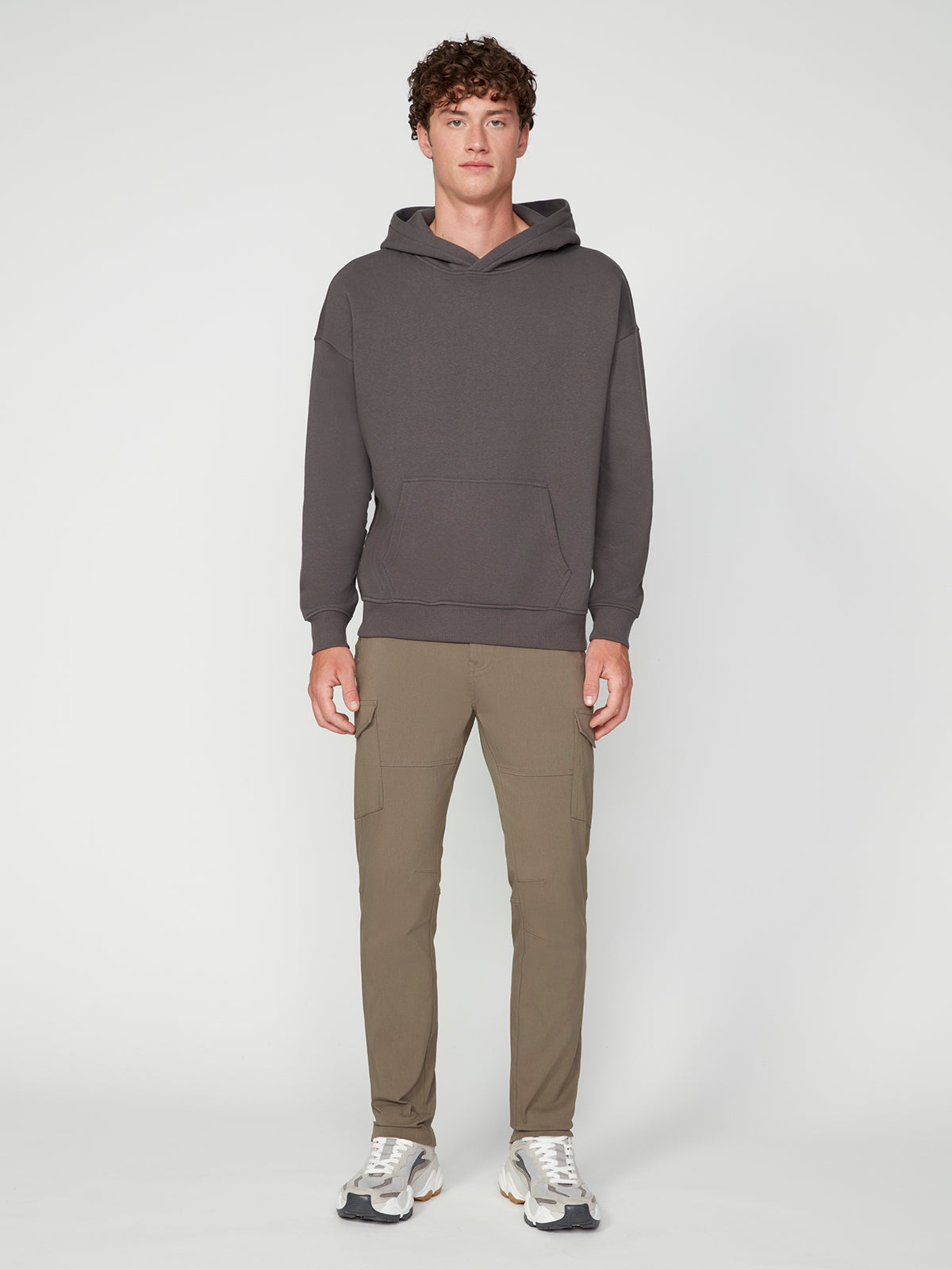A young man stands facing forward against a light gray background, wearing a PROJEK RAW MENS LONG SLEEVE CARBON PEACHED FLEECE HOODIE in dark gray, paired with light brown cargo pants and white sneakers.