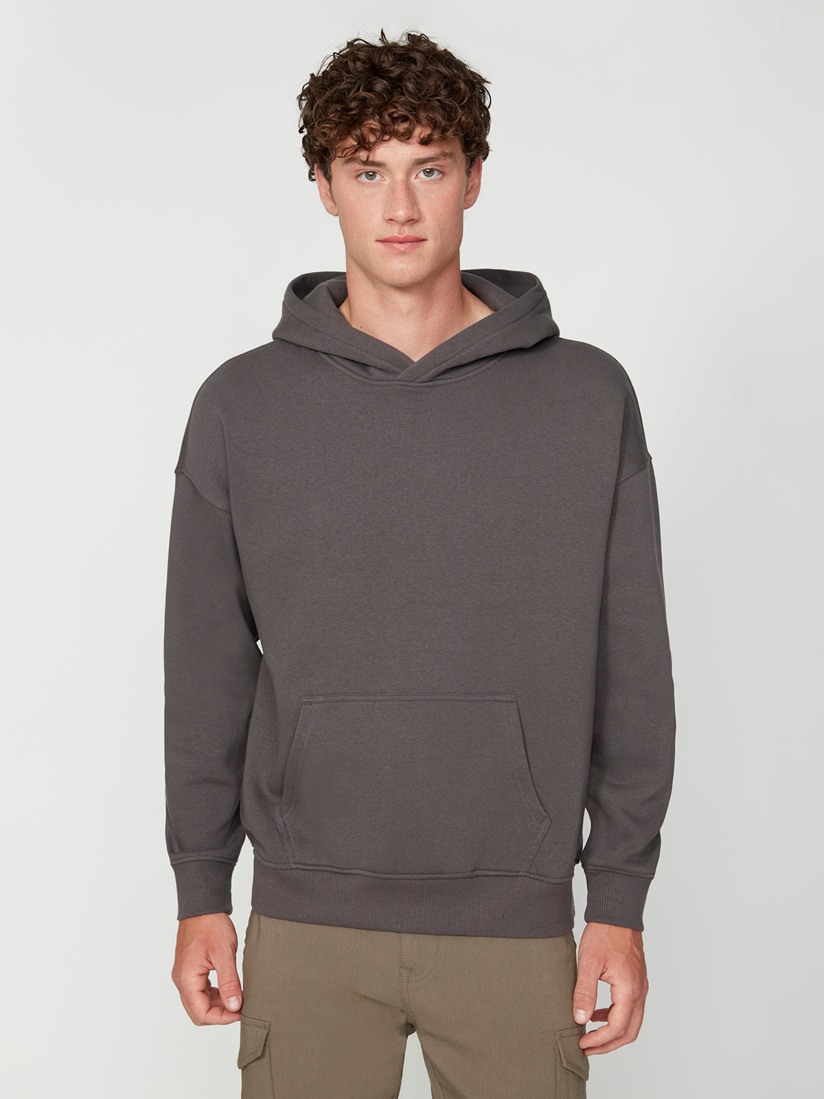 A young man with curly brown hair models the PROJEK RAW MENS LONG SLEEVE CARBON PEACHED FLEECE HOODIE in dark gray, featuring a front pocket, paired with tan cargo pants, against a light gray background.