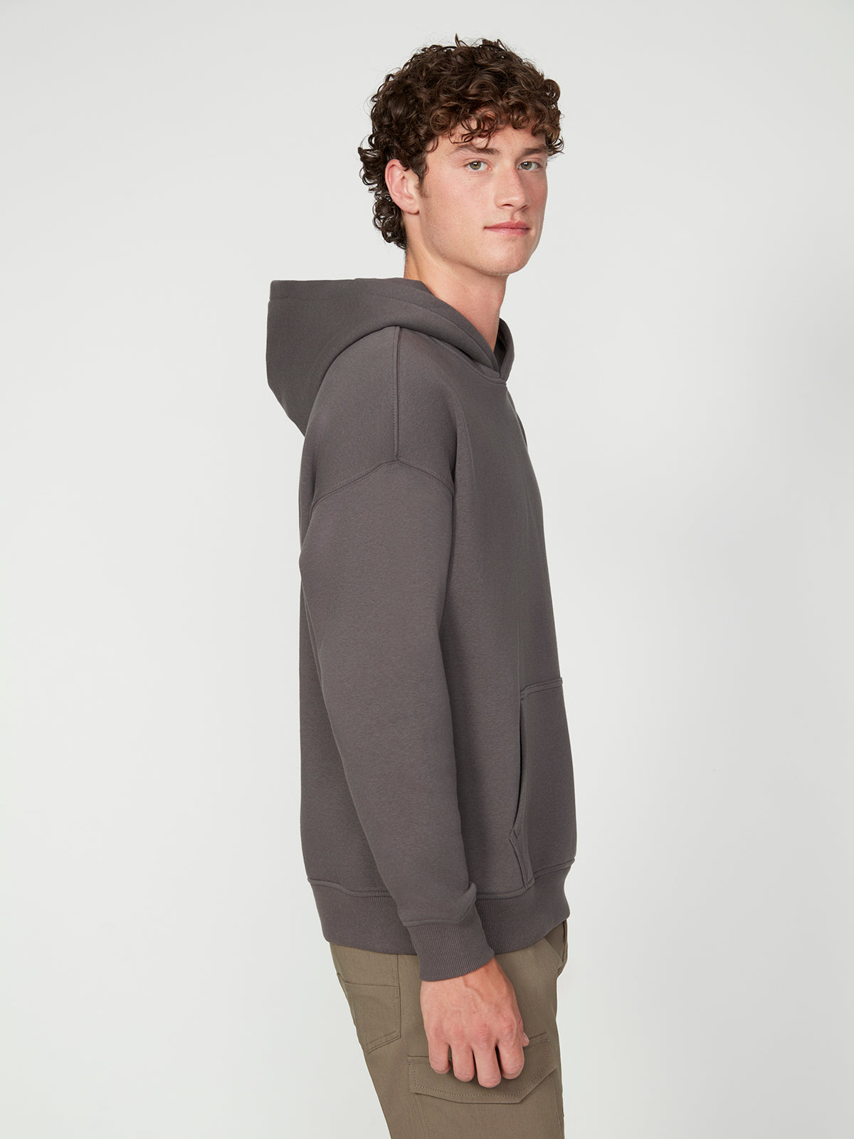 A young man with curly hair models the PROJEK RAW MENS LONG SLEEVE CARBON PEACHED FLEECE HOODIE in gray and khaki pants, standing sideways and looking toward the camera against a plain white background.