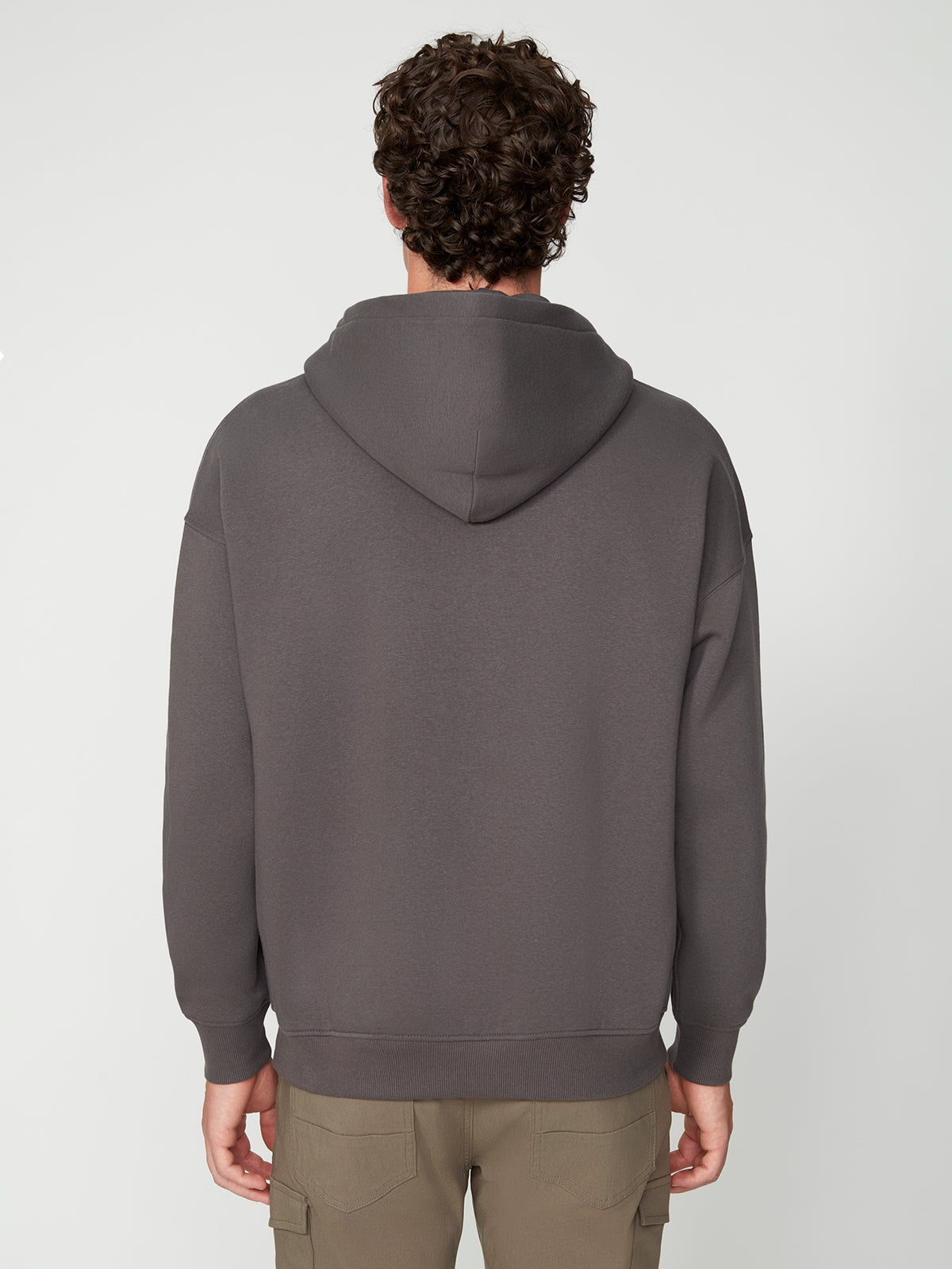 A person with curly hair is seen from the back, wearing a PROJEK RAW MENS LONG SLEEVE CARBON PEACHED FLEECE HOODIE and light brown cargo pants, standing against a plain light background.