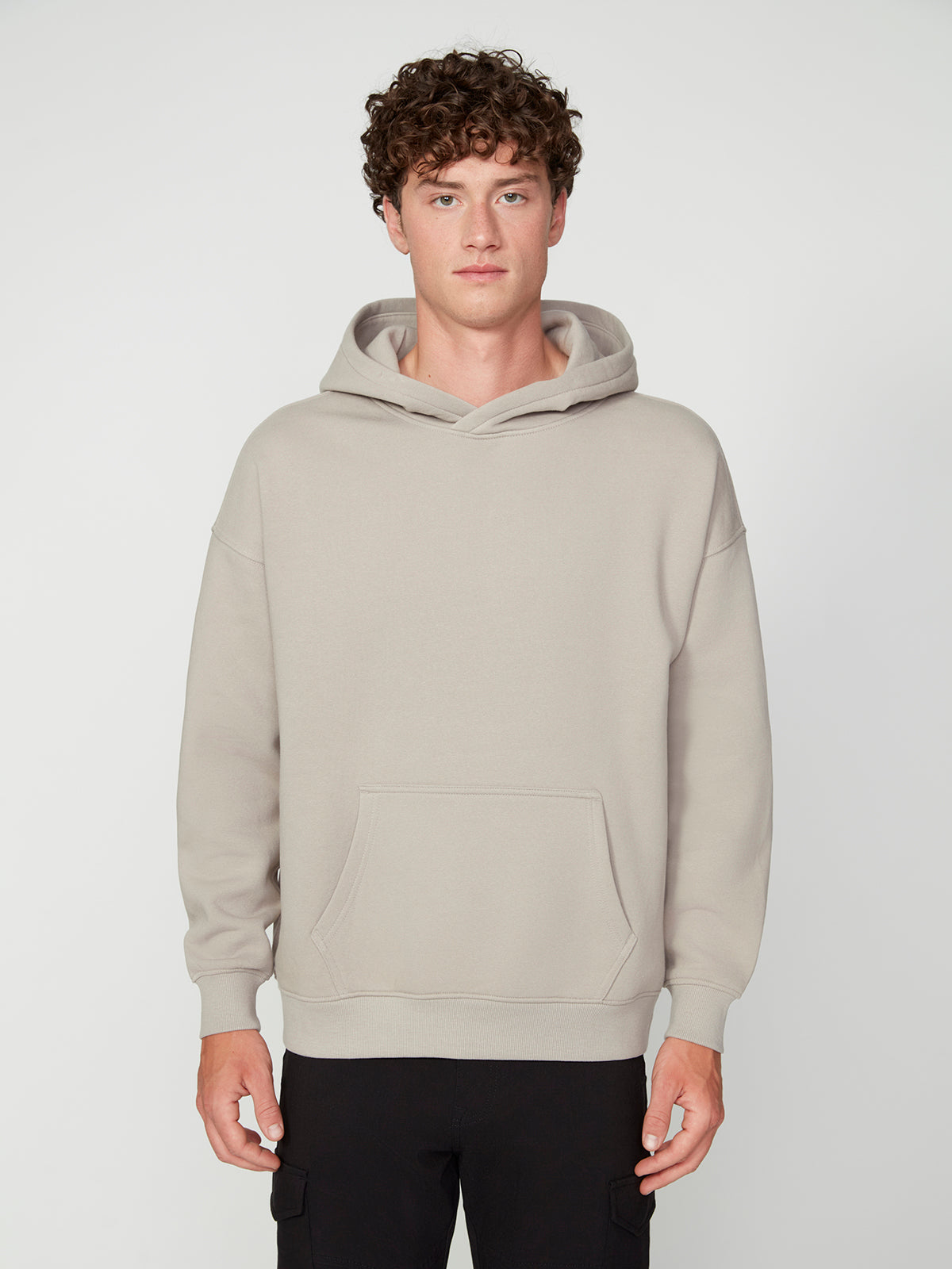 A young person with curly brown hair wears the PROJEK RAW MENS LONG SLEEVE CARBON PEACHED FLEECE HOODIE in light beige and black pants, standing facing forward against a plain, light background.