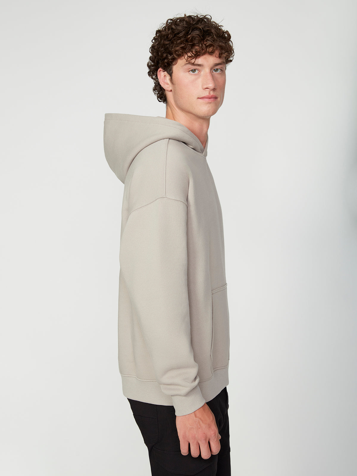 A young man with curly hair models the PROJEK RAW MENS LONG SLEEVE CARBON PEACHED FLEECE HOODIE in light gray and black pants, standing sideways and facing the camera against a plain white background.
