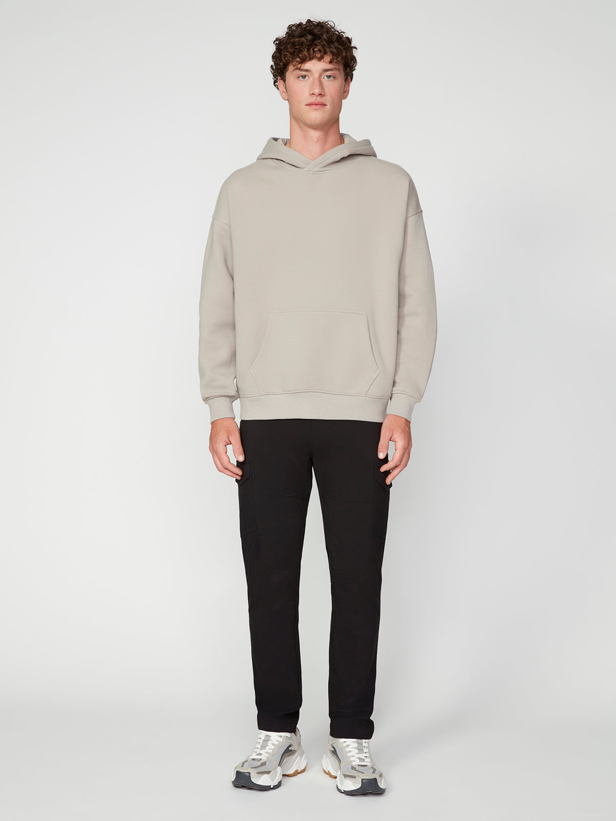 A young man faces forward in a plain light setting, wearing the PROJEK RAW MENS LONG SLEEVE CARBON PEACHED FLEECE HOODIE in light beige, paired with black pants and white sneakers.