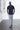 A man wears PROJEK RAW’s SOLID BUNGEE LOOSE FIT CHINO PANT in light gray, a navy short-sleeve button-up shirt, and navy slip-on shoes, standing with hands in pockets in a bright minimalist room with white walls and a concrete floor.