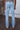A person stands indoors facing away from the camera, wearing PROJEK RAW’s HIGH RISE SKINNY JEAN in light blue with tan shoes. The urban streetwear setting includes a white brick wall with exposed brick and pipes.