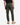 A person is standing facing away from the camera against a plain white background, wearing PROJEK RAW’s SOLID BUNGEE PULL ON JOGGER in olive green with an adjustable waist, paired with a black shirt and tan suede boots.