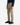 A person stands slightly turned against a plain background, hands relaxed by their sides and wearing PROJEK RAW’s SOLID BUNGEE 5-POCKET RELAXED FIT PANTS in beige—a contemporary essential crafted from a premium stretch blend—paired effortlessly with a blue plaid shirt and black sneakers.