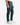 A person dressed in PROJEK RAW’s CAVALRY TWILL BUNGEE CHINO PANTS in teal, paired with a crisp white shirt and white sneakers, stands against a simple light background—an effortless approach to modern office style.