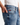 A person wearing the PROJEK RAW BARU CLASSIC STRAIGHT FIT DENIM in light blue, vintage-inspired wash stands with one hand in their pocket. The straight-leg jeans, featuring a red tag above the coin pocket, are effortlessly paired with a white ribbed tank top.