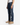 A person stands against a plain white background, with only the lower half of their body visible, wearing PROJEK RAW’s BARU CLASSIC STRAIGHT FIT DENIM in indigo, paired with a white tank top and tan suede shoes.