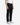 A person is wearing BARU CLASSIC TWILL PEACHED DENIM by PROJEK RAW—black straight-leg jeans—along with a white sleeveless shirt and white sneakers, standing against a plain white background. Only the lower torso and legs are visible.