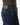 A close-up side view of a person wearing PROJEK RAW’s ATLAS ATHLETIC FIT DENIM in a midnight blue wash with a tapered leg, featuring a leather patch on the waistband and a small red label above the back pocket, paired with a brown top.