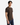 A man with straight black hair, dressed in the PROJEK RAW KNIT CORD RELAXED FIT CREWNECK in dark brown and blue jeans, stands with one hand in his pocket against a plain white background.