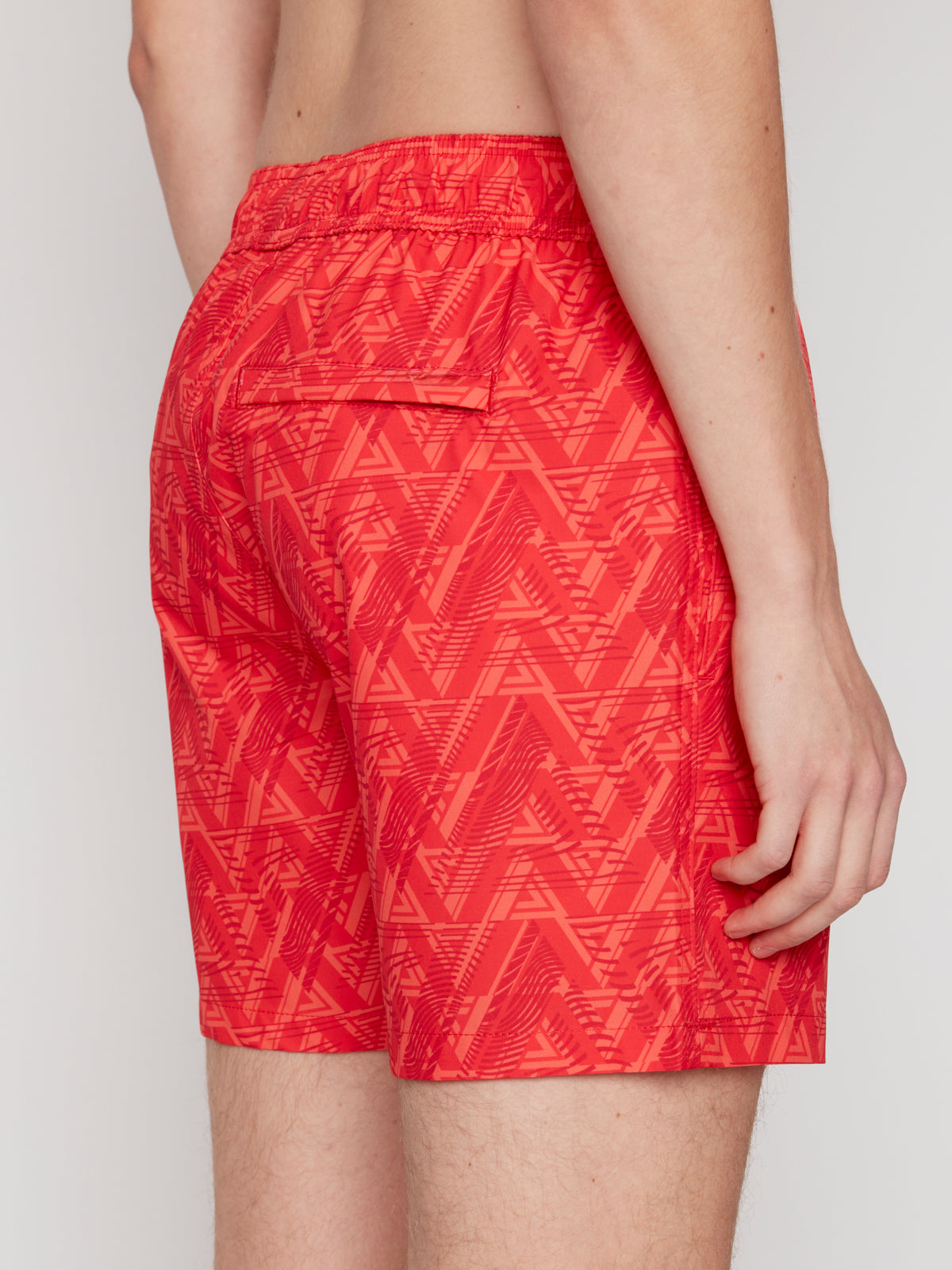 A person is shown from the waist down, side and back view, wearing Projek Raw BRYSON-CORAL swim trunks featuring a geometric red pattern, elastic waistband, and a back flap pocket.