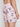 A person wearing Projek Raws PATRICK-PINK swim trunks, featuring a light pink tropical print with flamingos, palm trees, small islands, an elastic waistband, and a zippered back pocket.