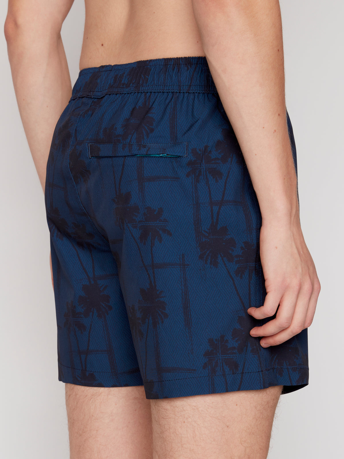 A person is shown from the waist down wearing Projek Raw BRUNO-NAVY swim shorts featuring a dark palm tree pattern, elastic waistband, and back pocket. Their left arm rests relaxed at their side.