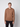 A young man faces forward in a PROJEK RAW Mens Long Sleeve Hoodie with Kangaroo Pocket, light brown in color, paired with black pants. The background is plain and light-colored.
