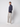 A man stands against a plain light background, wearing a navy turtleneck sweater, PROJEK RAW Mens Relaxed 5-Pocket Pant in light gray, and white sneakers, with his hands in his pockets.