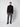 A man stands against a light background, wearing a black turtleneck, PROJEK RAW Men’s Relaxed Chino Pant in gray-green, and black shoes. He faces forward with a neutral expression and relaxed posture.