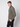 A young man with short light brown hair wears the PROJEK RAW Mens Casual Dress Shirt with Logo Detail—an olive green button-up—paired with black pants, standing against a plain light gray background and looking at the camera.