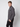 A young man with short light brown hair wears the PROJEK RAW Mens Casual Dress Shirt with Logo Detail in dark gray and black pants, standing sideways with one hand in his pocket against a plain light background for a modern, effortless look.