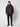 A man faces forward with a neutral expression, wearing a black PROJEK RAW Mens Textured Ripstop Ecofill Quilted Parka with Hood over a red zip-up sweater, paired with black pants and black boots against a plain background.