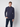 A young man with short light brown hair stands against a plain light gray background, wearing the PROJEK RAW Mens 24/7 Button-up Shirt in dark blue and dark pants, one hand in his pocket. The shirt is travel-friendly and wrinkle-resistant.