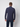A man faces away from the camera, wearing PROJEK RAWs Mens 24/7 Button-up Shirt in dark navy with dark pants, set against a plain light background. The shirt is travel-friendly and wrinkle-resistant.