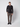 A young man stands against a plain background, wearing the PROJEK RAW Mens 24/7 Button-up Shirt in black with brown plaid trousers and black shoes, one hand in his pocket and a neutral expression.
