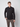 A person stands against a plain light background, wearing the PROJEK RAW Mens 24/7 Button-up Shirt in black with long sleeves and plaid brown trousers, one hand in their pocket and a neutral expression.