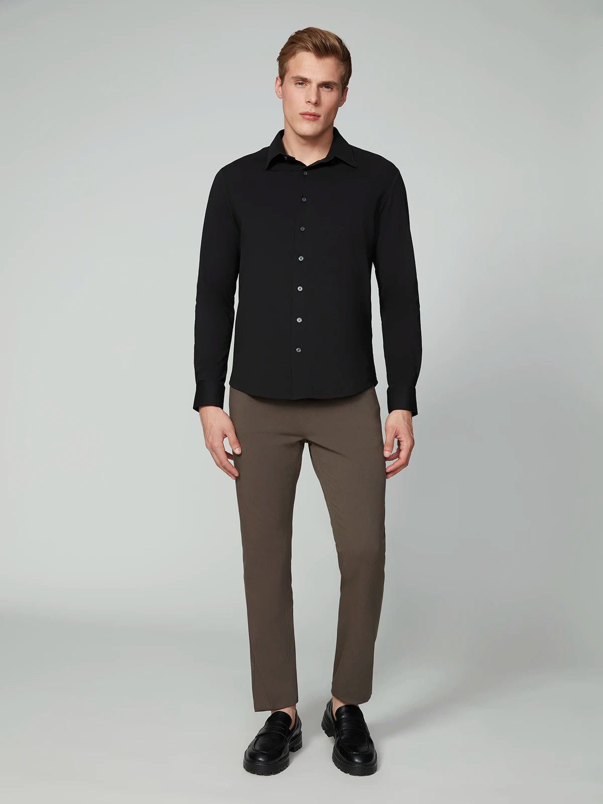 A man faces forward against a plain light gray background, wearing the PROJEK RAW MENS 24/7 BUTTON-UP KNIT SHIRT in black, taupe slim-fit pants, and black shoes.