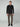 A man faces forward against a plain light gray background, wearing the PROJEK RAW MENS 24/7 BUTTON-UP KNIT SHIRT in black, taupe slim-fit pants, and black shoes.