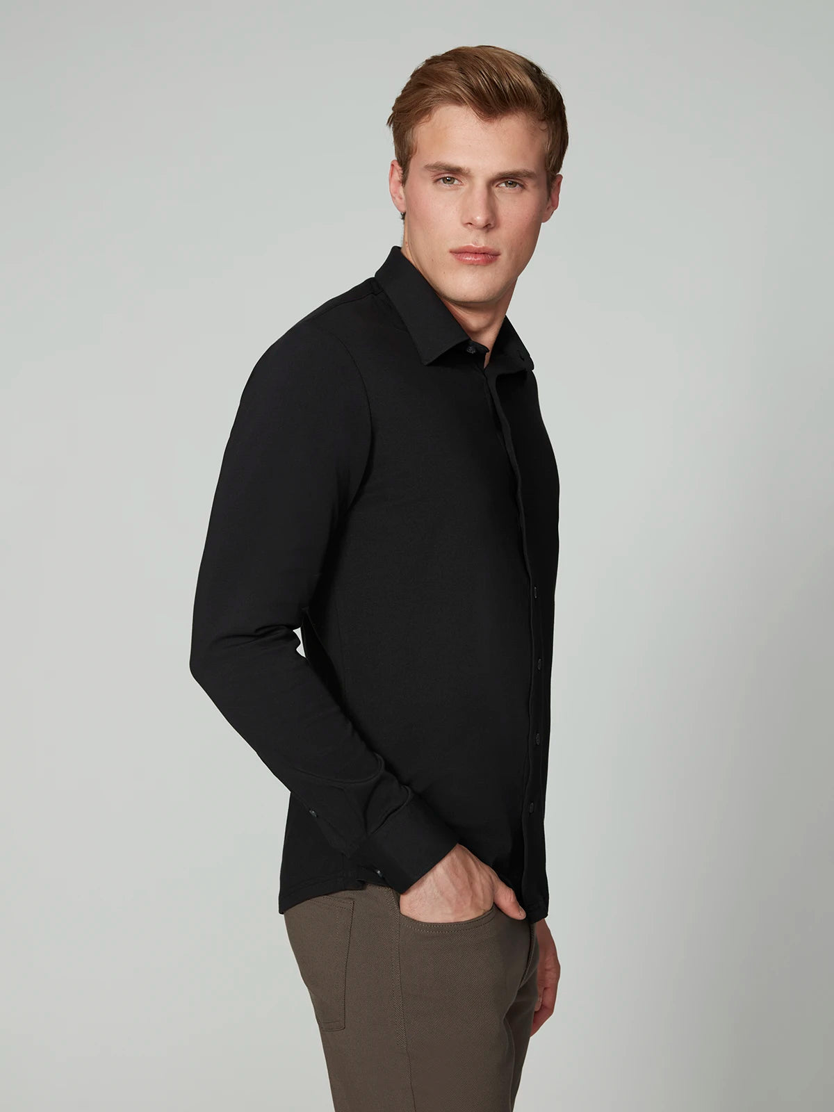 A young man with short blonde hair wears the PROJEK RAW MENS 24/7 BUTTON-UP KNIT SHIRT and brown pants, standing with one hand in his pocket against a plain light gray background.
