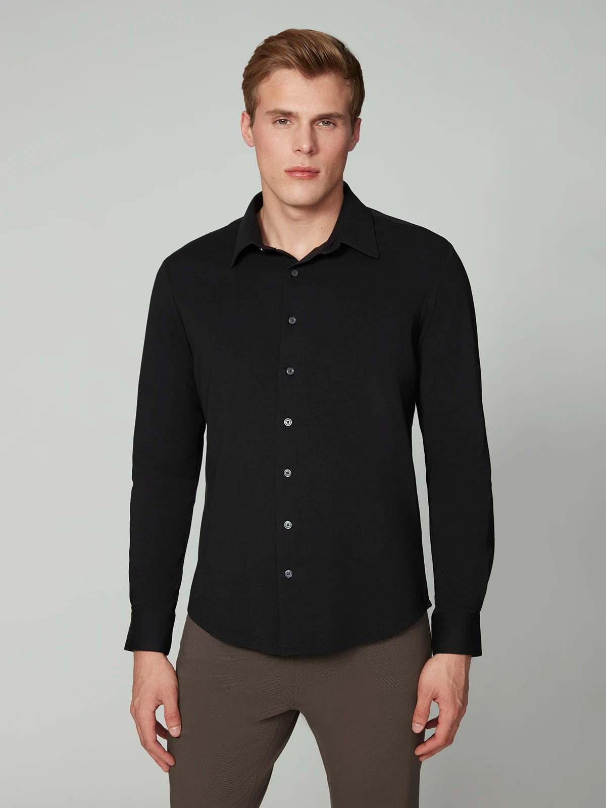 A man with short blond hair wears the PROJEK RAW MENS 24/7 BUTTON-UP KNIT SHIRT in black with brown pants, standing against a plain light gray background.
