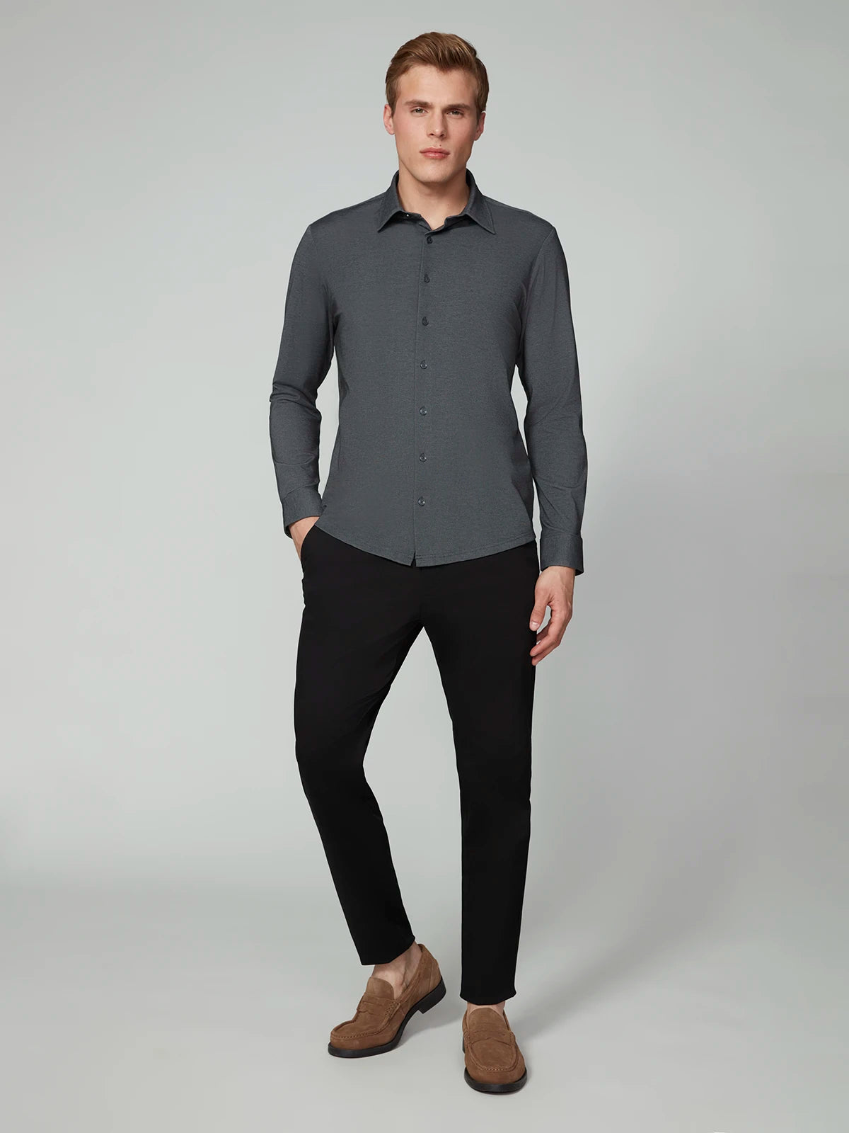 A man with short blond hair and a neutral expression stands against a plain background, wearing the PROJEK RAW MENS 24/7 BUTTON-UP KNIT SHIRT in dark gray, paired with black pants and light brown suede loafers.