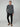 A man with short blond hair and a neutral expression stands against a plain background, wearing the PROJEK RAW MENS 24/7 BUTTON-UP KNIT SHIRT in dark gray, paired with black pants and light brown suede loafers.