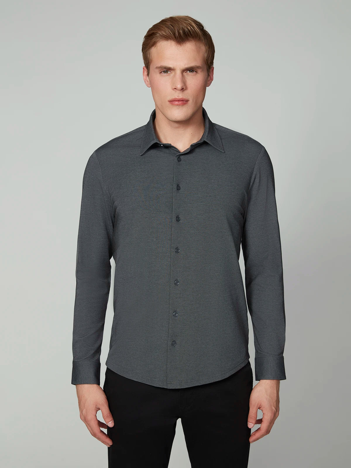 A man with short blonde hair models the PROJEK RAW MENS 24/7 BUTTON-UP KNIT SHIRT in dark gray with black pants, standing against a light gray background and looking directly at the camera with a neutral expression.