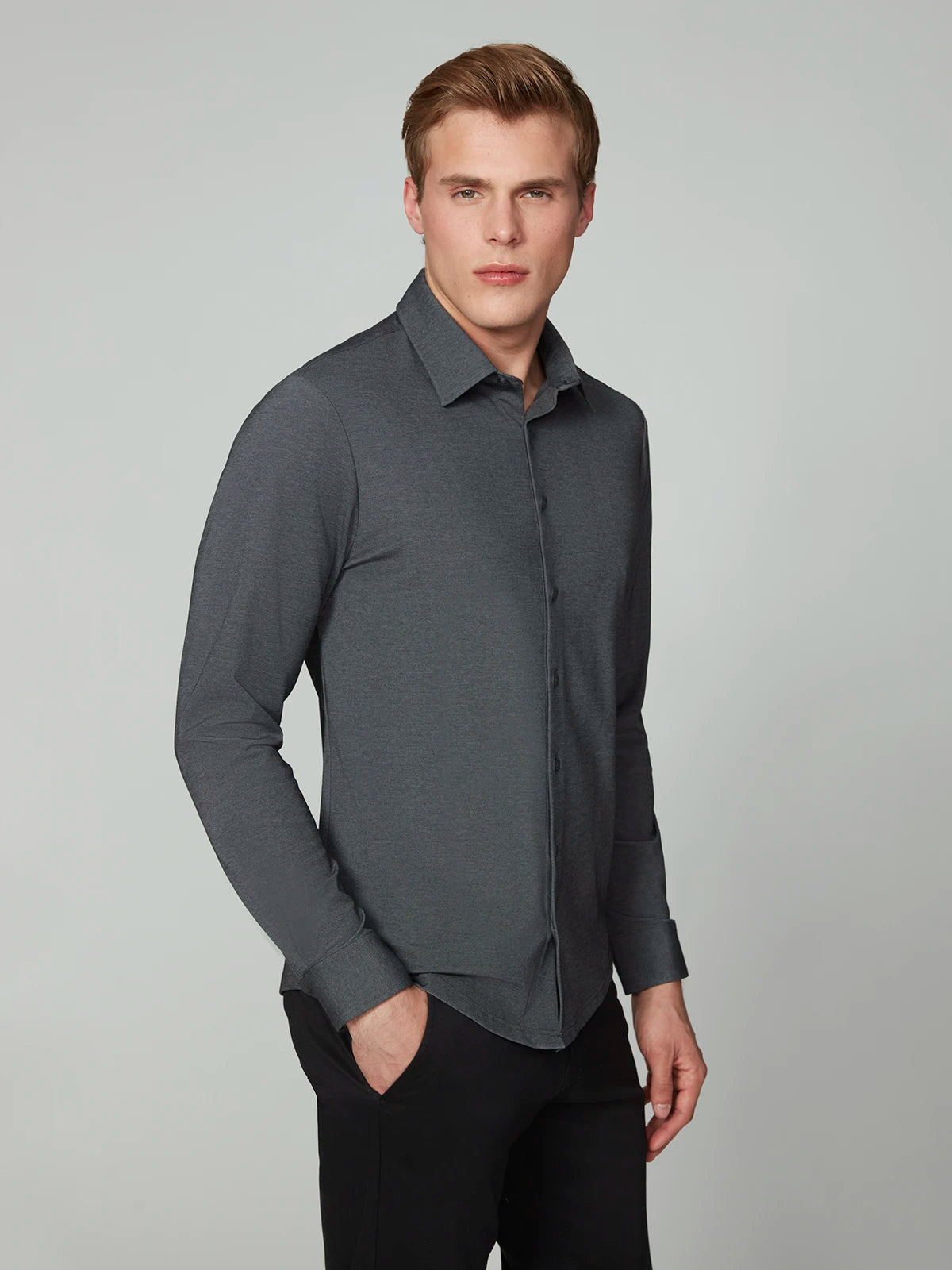 A man with short light brown hair wears the PROJEK RAW MENS 24/7 BUTTON-UP KNIT SHIRT and black pants, standing facing forward with one hand in his pocket against a light gray background.