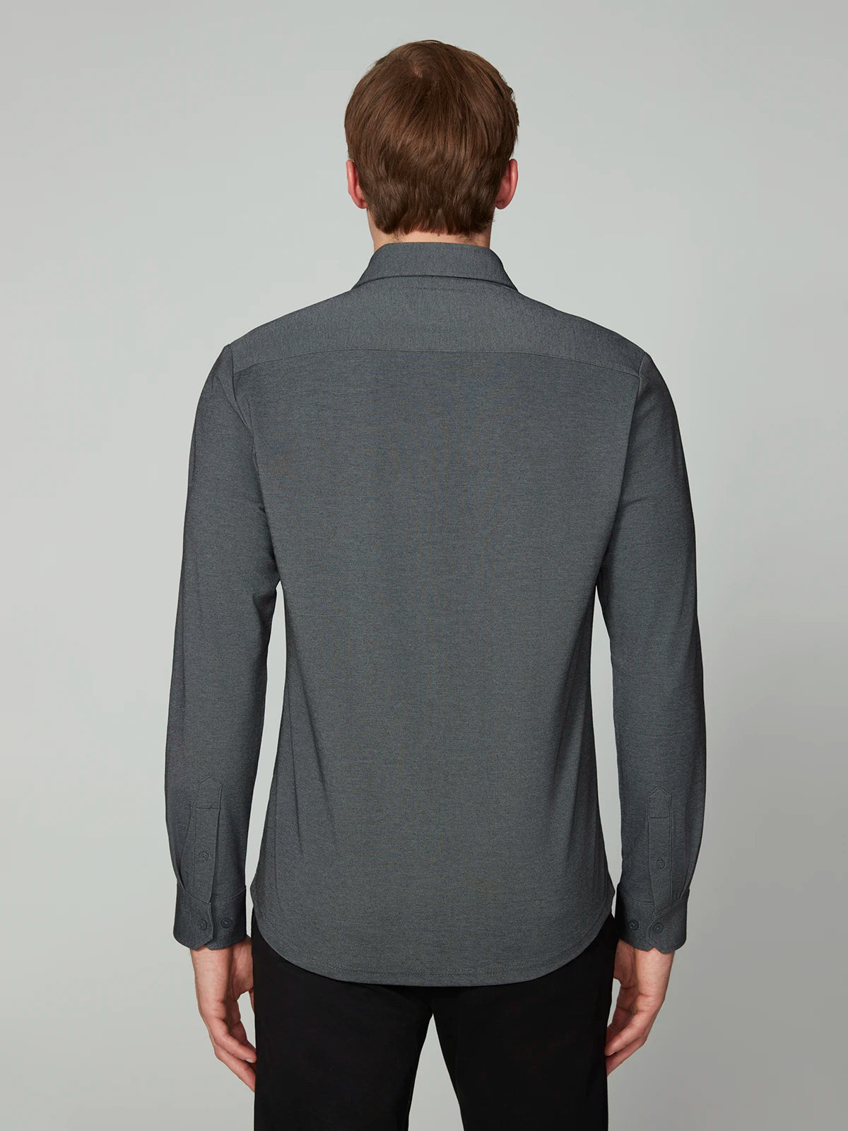 A person with light brown hair is shown from behind, wearing the PROJEK RAW MENS 24/7 BUTTON-UP KNIT SHIRT in charcoal gray with black pants, set against a plain light gray background.