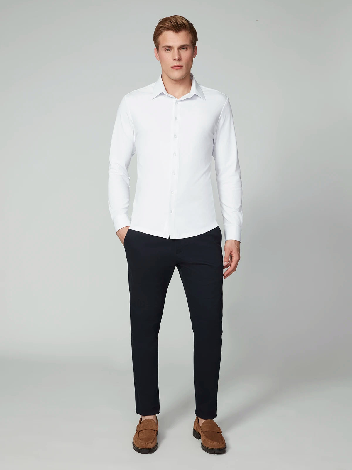 A man faces forward against a plain light background, wearing the PROJEK RAW MENS 24/7 BUTTON-UP KNIT SHIRT in white, dark pants, and brown loafers, with his left hand in his pocket.